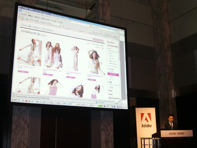 Adobe Scene7の導入について紹介する，株式会社ピーチ・ジョン執行役員 守安智氏。導入動機について，ユーザからの多様なニーズへの対応，リッチな体験の提供，カタログのアウトソース費の削減など，さまざまなメリットについて述べた。